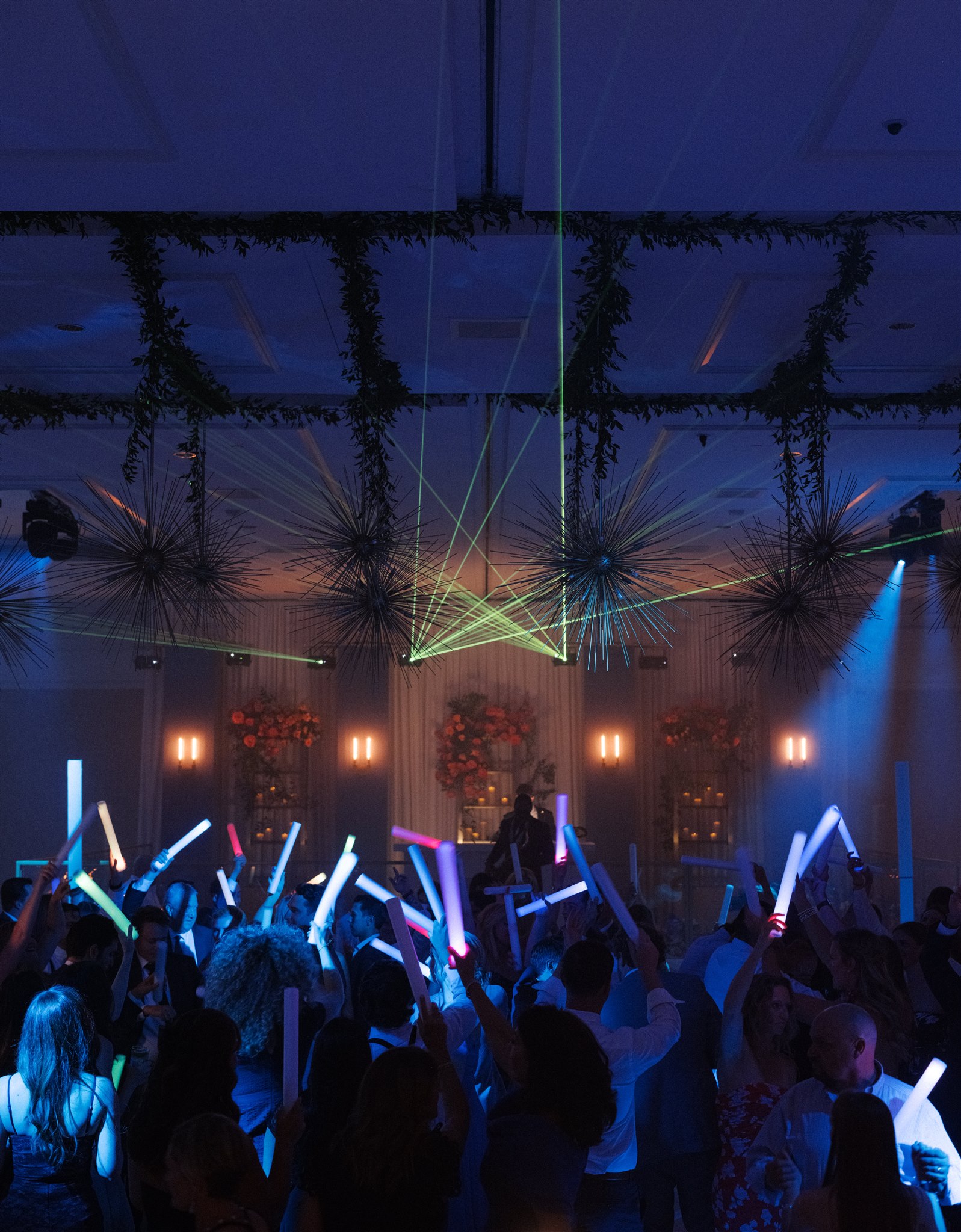 Wedding guests holding light-up foam props dance under a laser display at a late night wedding reception.