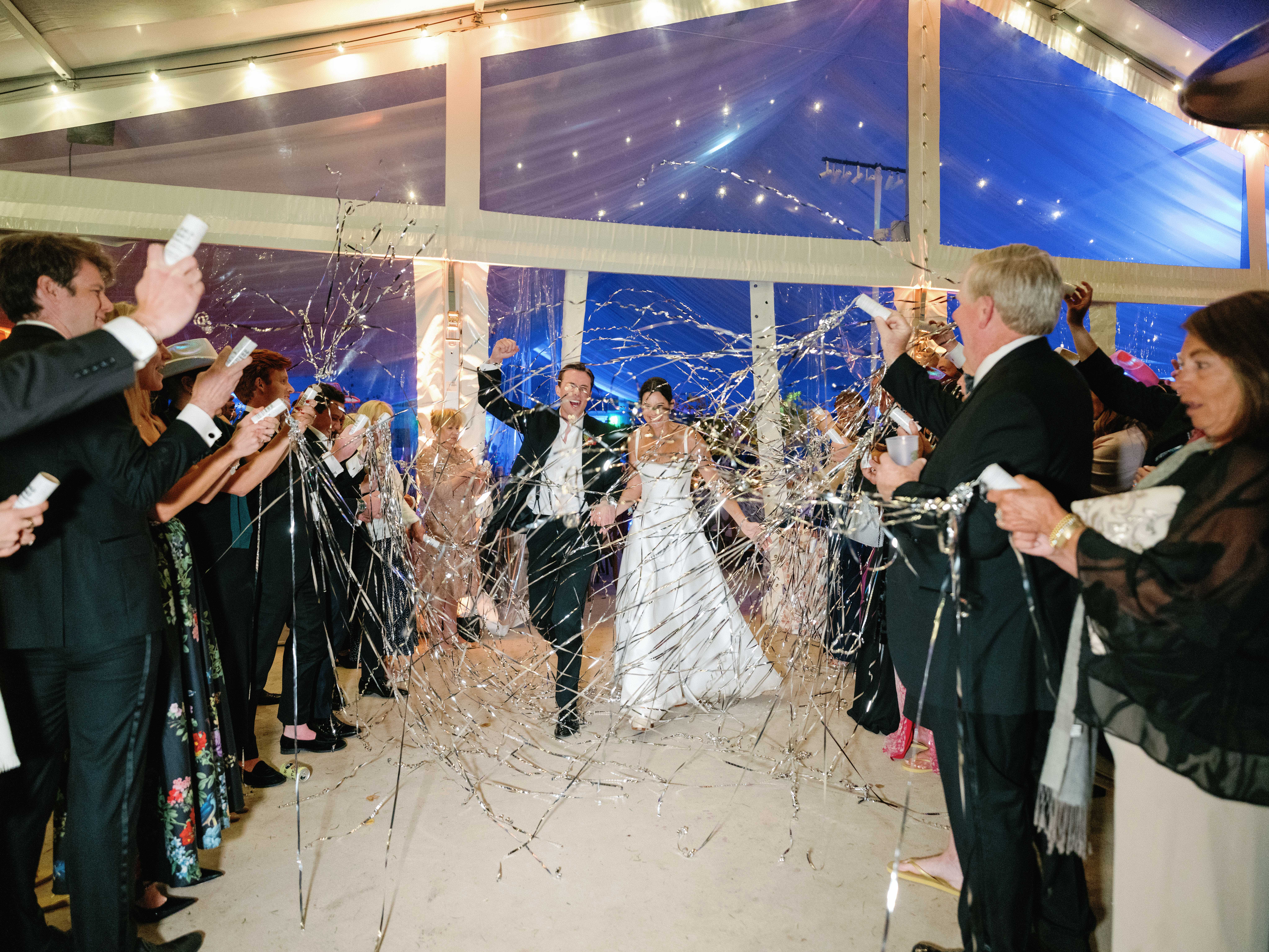 A bride and groom hold hands as their late night guests send them off with silver streamers.