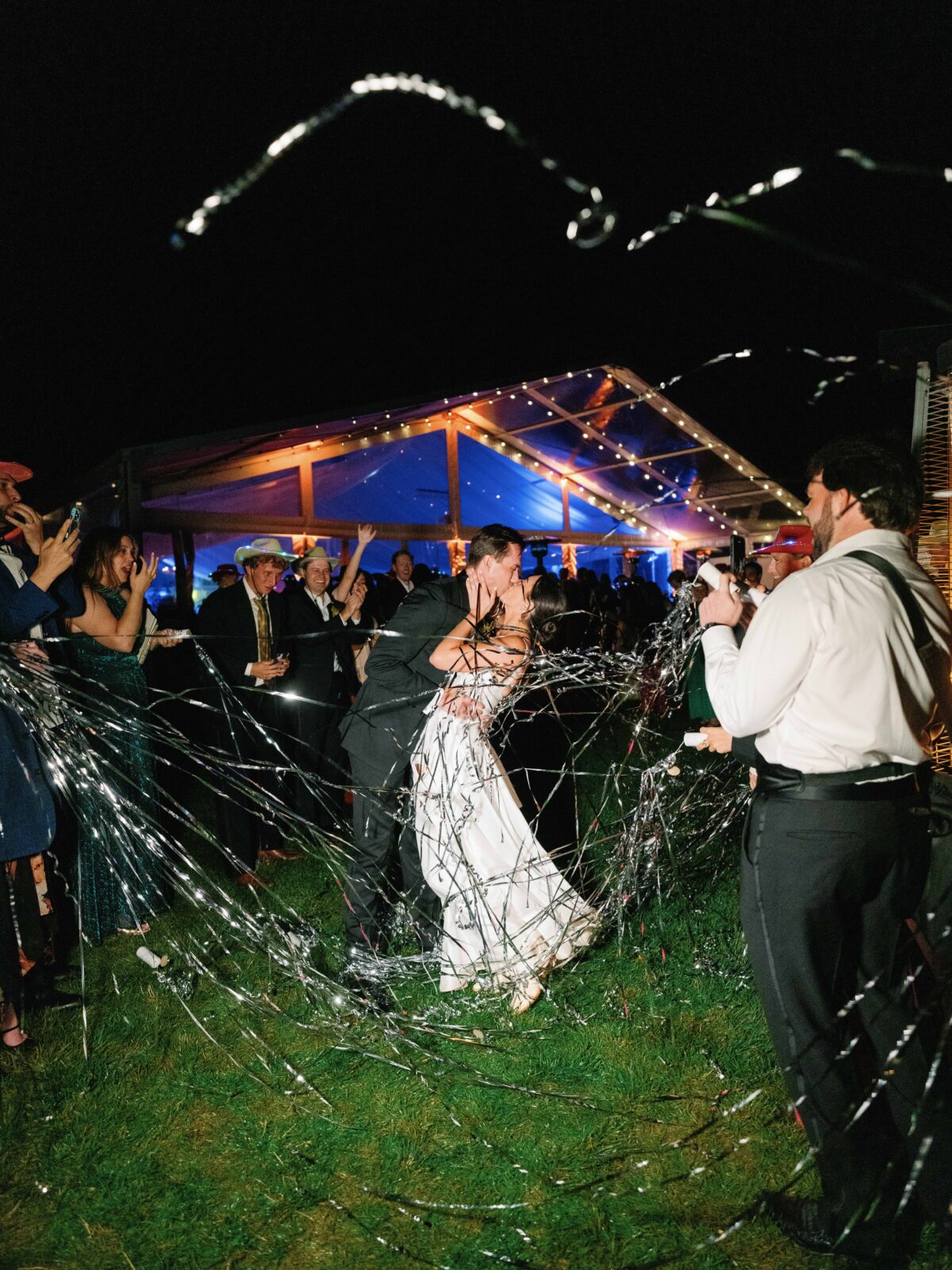A bride and groom share a kiss as their late night guests send them off with silver streamers.