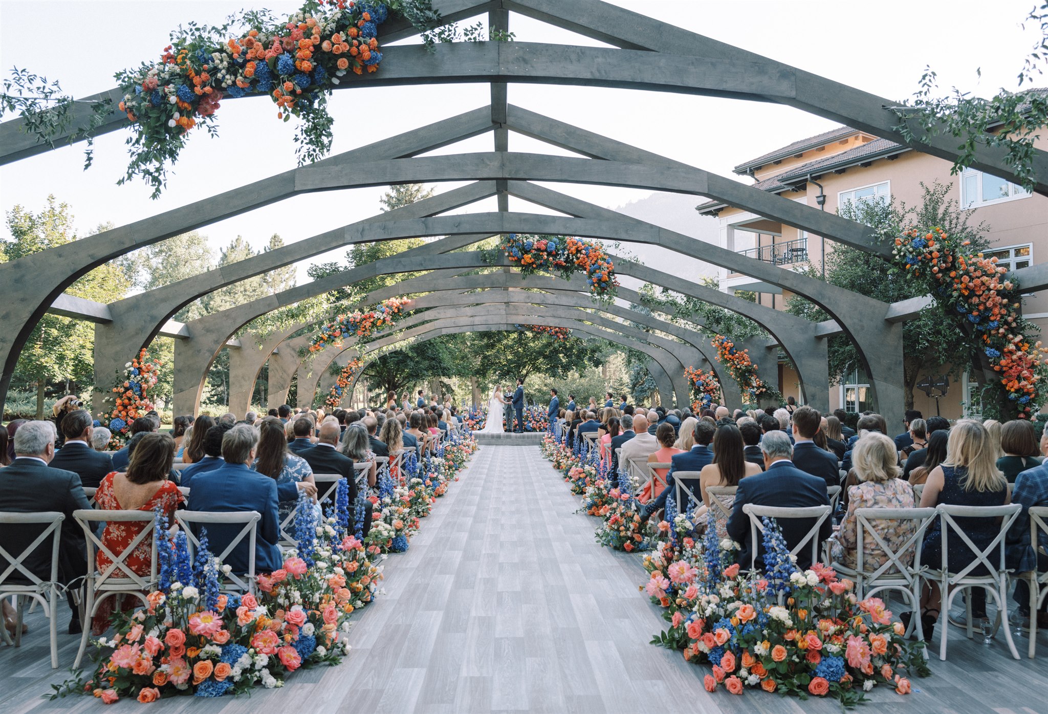 A large outdoor wedding beneath chapel-inspired wooden arches and coral and blue floral arrangements.