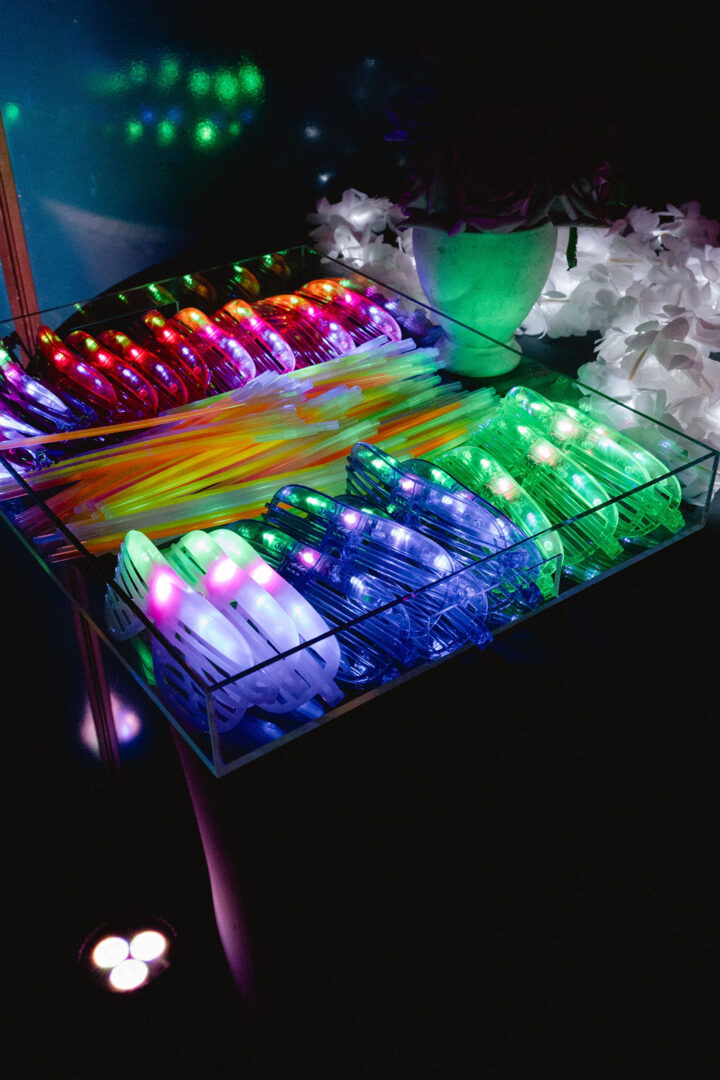 A clear acrylic tray of late night dance floor props that include light-up sunglasses, jelly rings, and glow sticks.