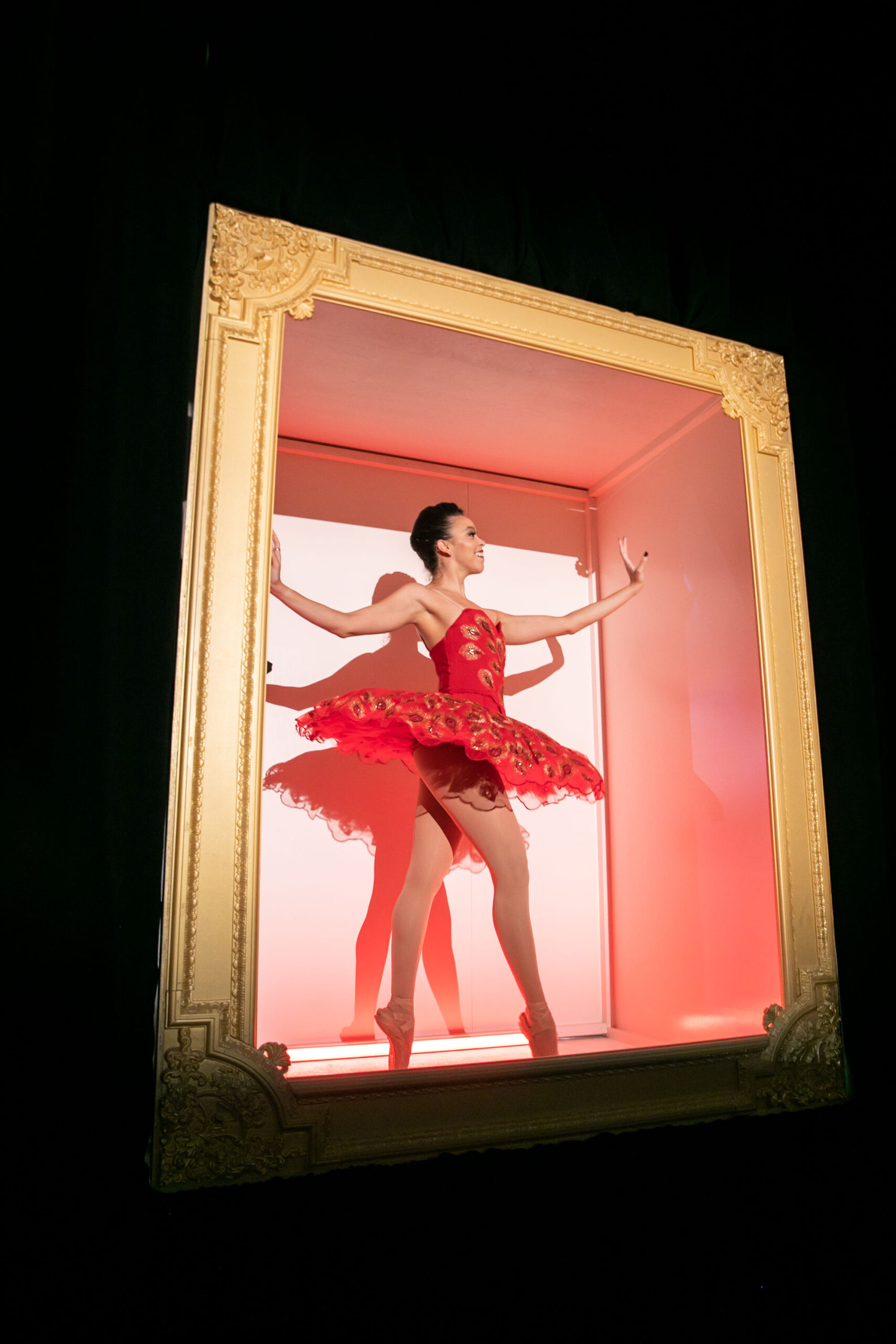 A ballerina in a red tutu dances in a large gold-framed shadowbox, providing entertainment at a wedding reception.