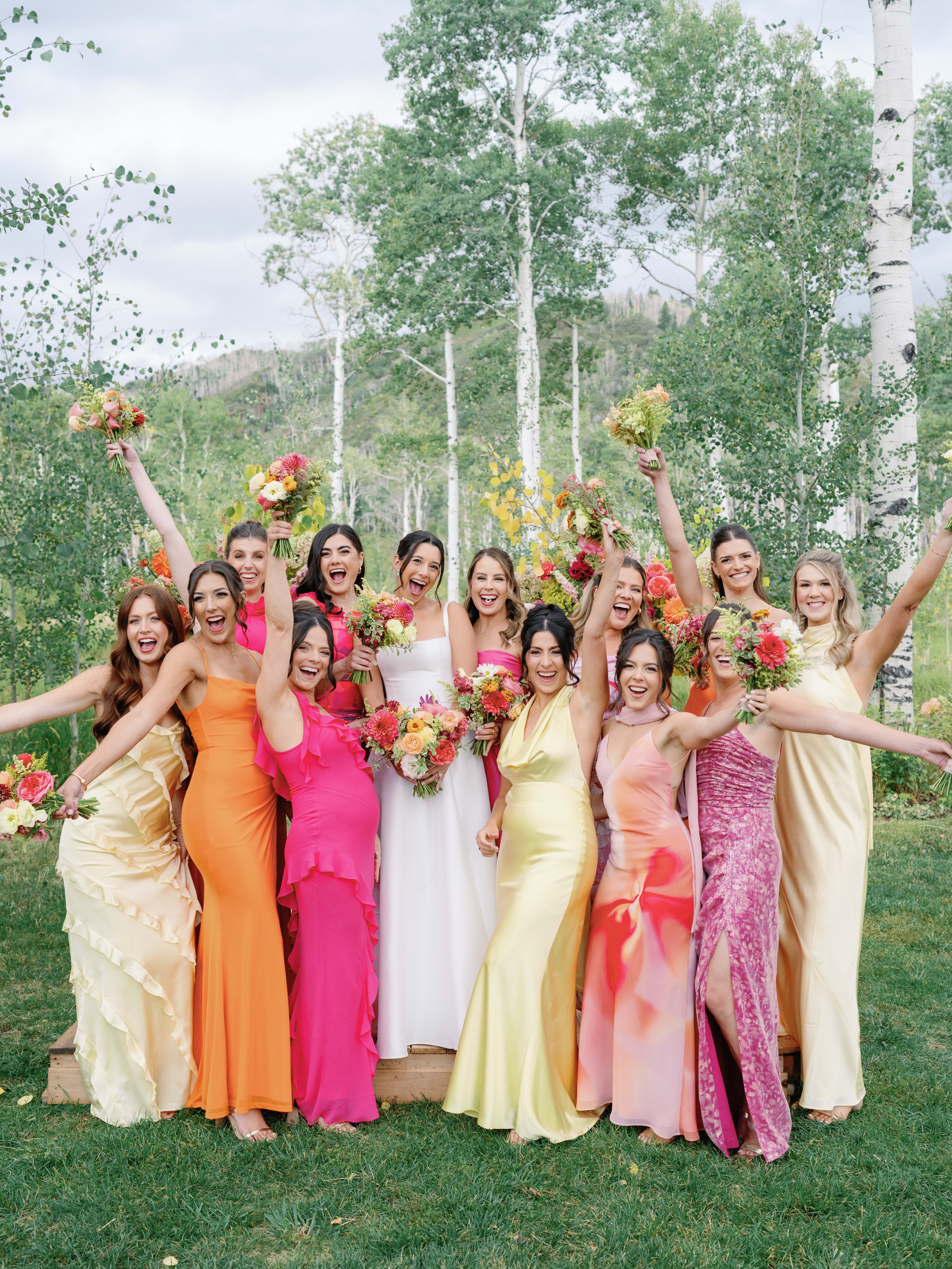 A dozen bridesmaids in varying shades of yellow, pink and orange surround a smiling bride in an Aspen grove.