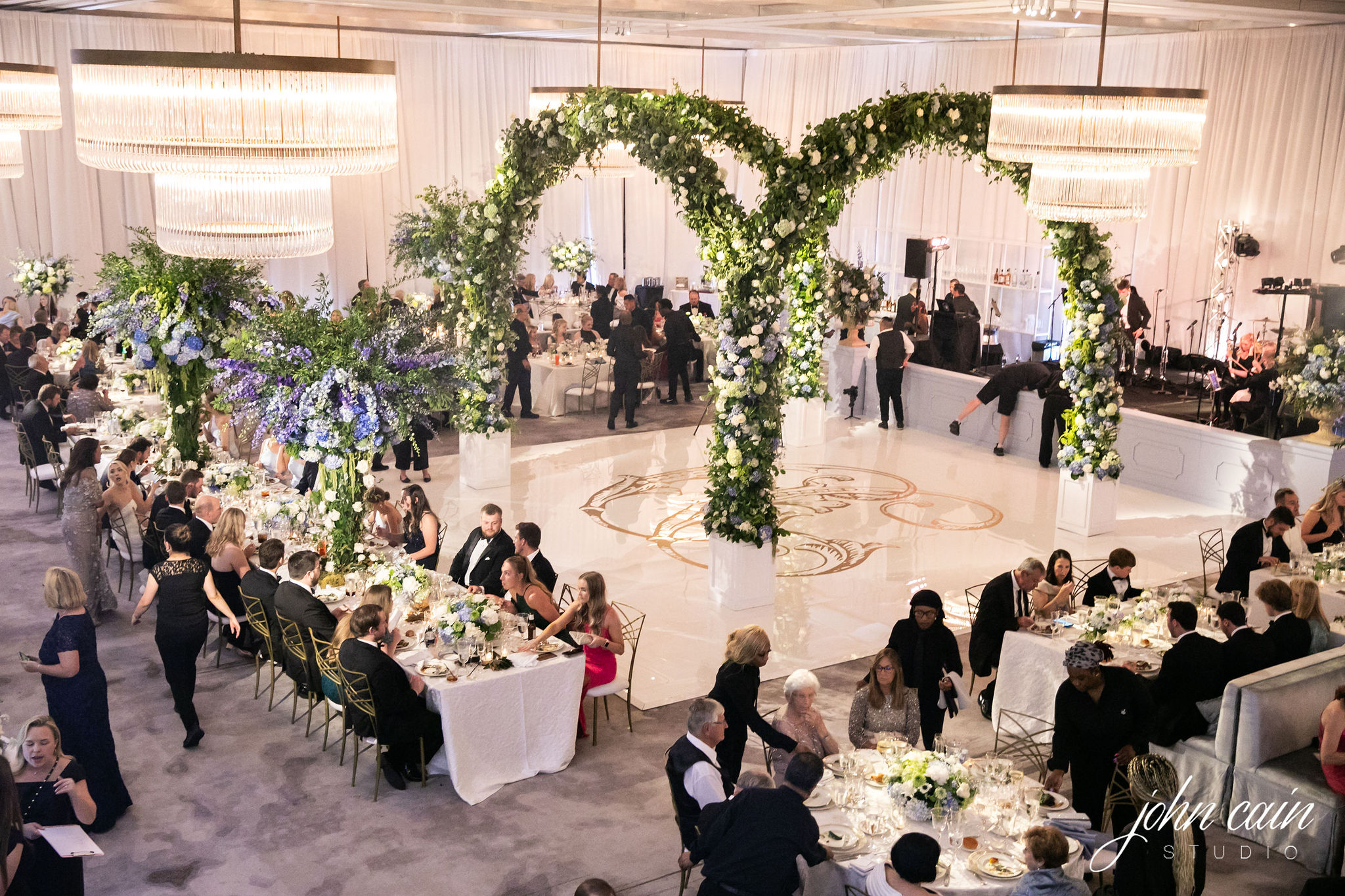 A wide room shot of a lavish wedding reception featuring a large four-sided greenery and floral arch in the middle of the dance floor.