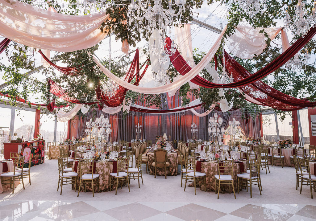 A grand, romantic tented wedding reception design featuring red and pink ceiling drapers, crystal chandeliers, and vintage accents.