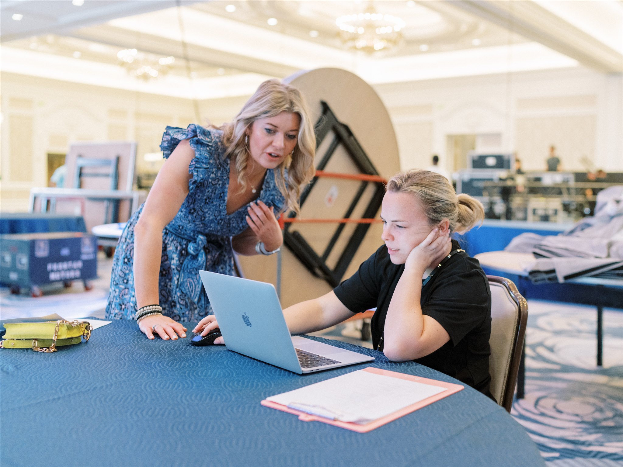 Two event planners work through last-minute details as a ballroom is set up behind them.