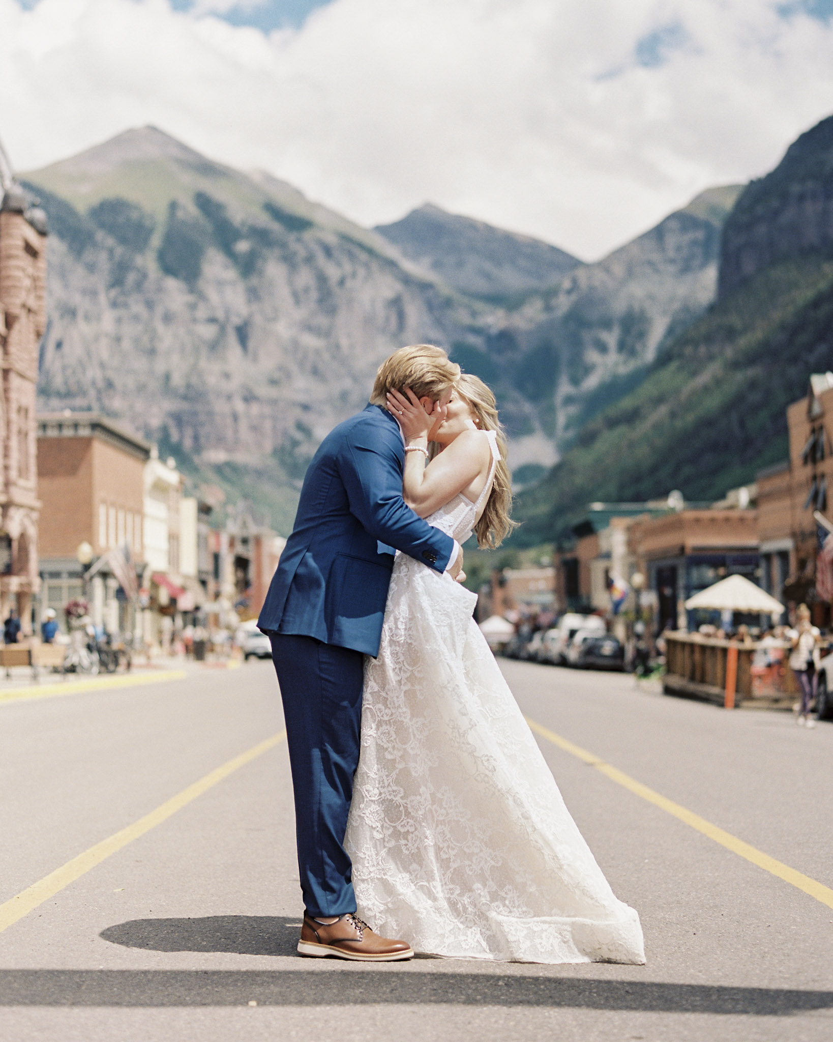 A bride and groom share a kiss in the middle of a small town street, against a mountainous backdrop